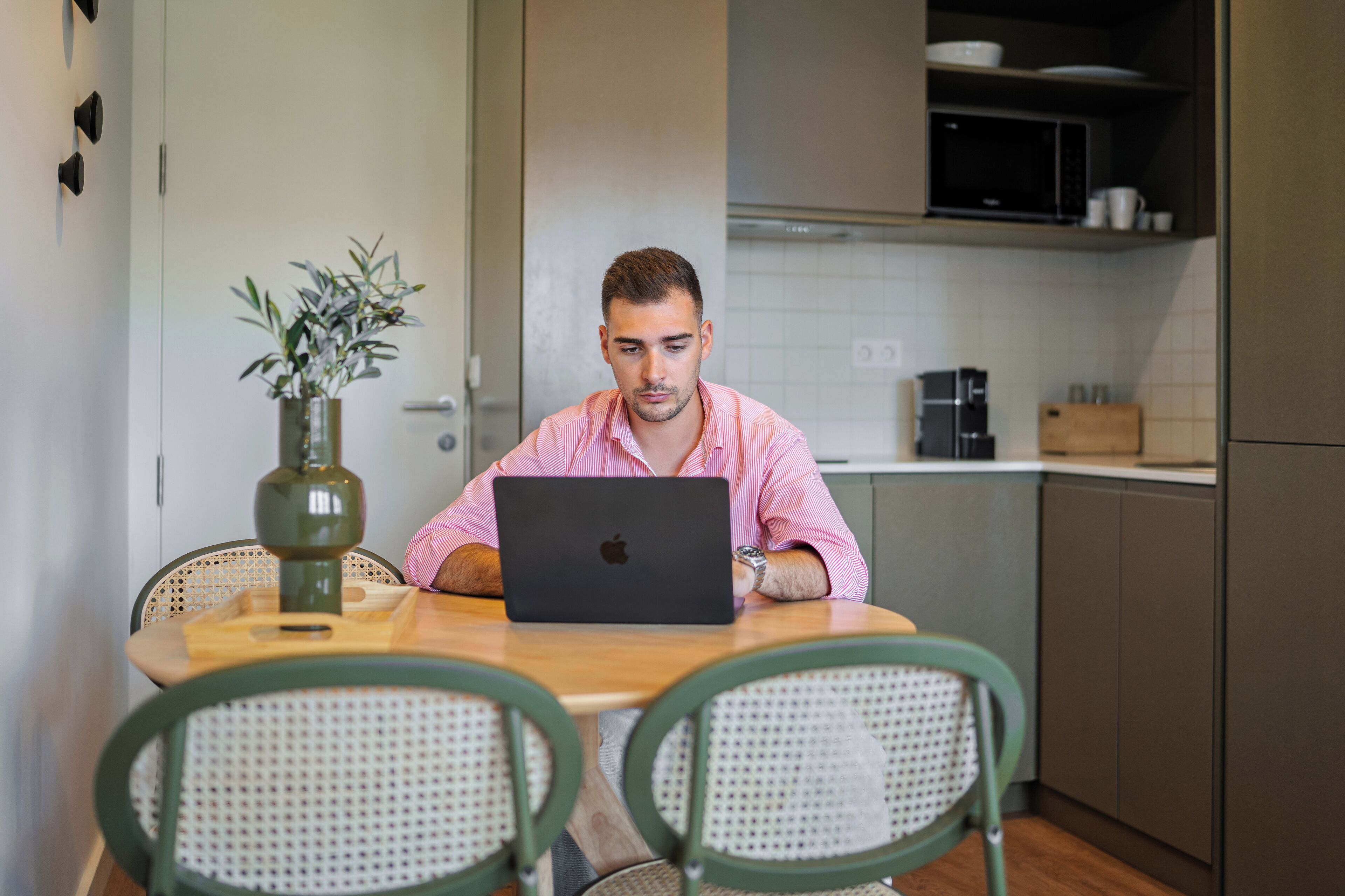 A person sitting at a wooden table with a laptop in a modern kitchen with wicker chairs, a vase with plants, and a microwave above the counter.