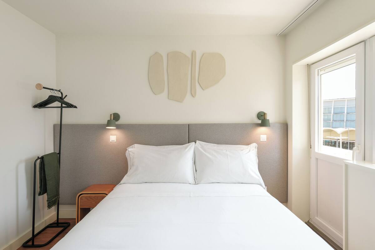 Habitación de hotel con cama, mesita de noche, lámpara, ventana y aplique de pared.