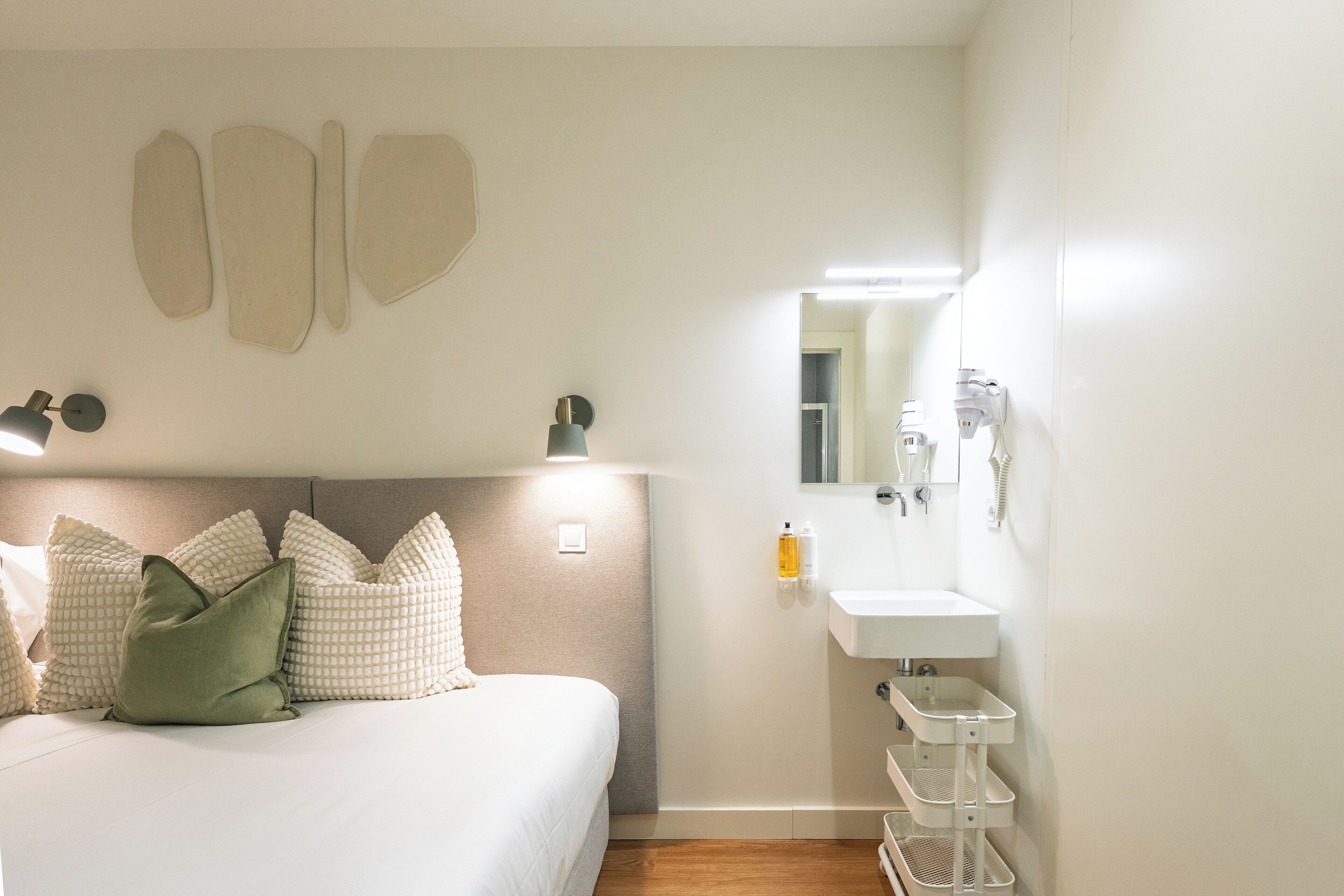 Een slaapkamer met een bed, kussens, een nachtkastje, een wandlamp, een spiegel en een badkamer met een wastafel en handdoekhouder.