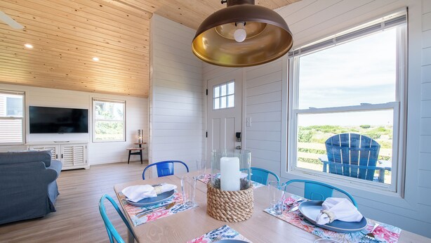 Un comedor con una mesa de madera dispuesta para cuatro, un ventanal con vista a la playa y un televisor en la sala.