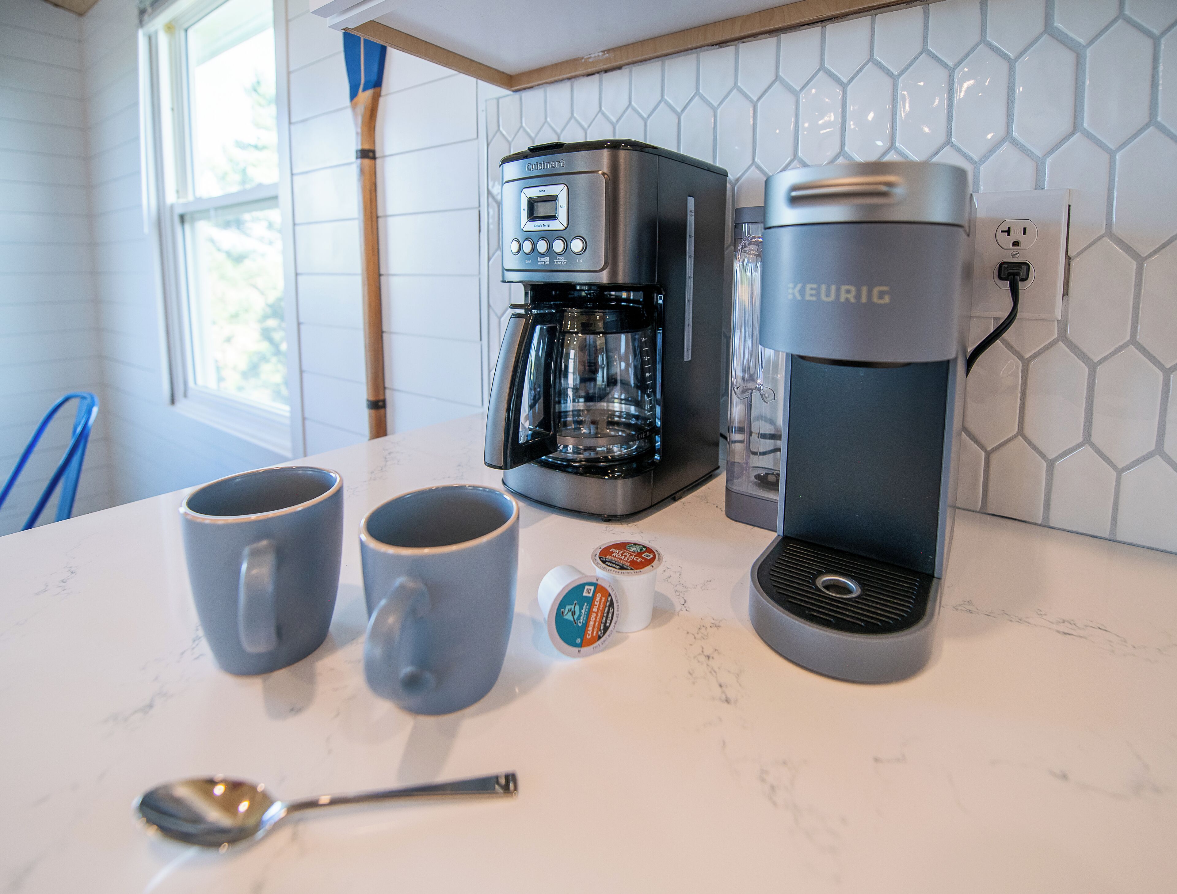 Una cocina con cafetera, dos tazas, una cuchara y una cafetera Keurig.