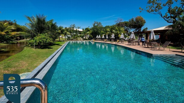 Piscina transparente com grade metálica, rodeada por espreguiçadeiras e guarda-sóis, e um pequeno lago à esquerda.
