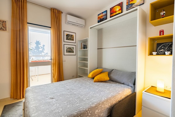 Un dormitorio con cama, armario, estante con objetos y ventana con cortinas.