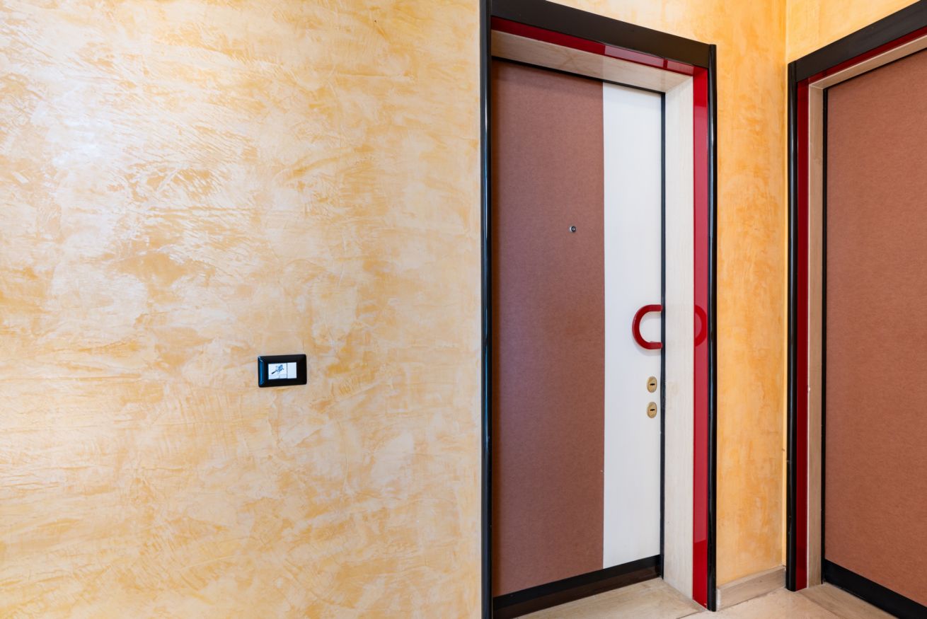 A hallway with a textured wall, a door with a red handle, and a light switch on the wall.