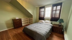 Une chambre avec un plafond en pente, un lit recouvert d’une couette grise, une table de chevet avec une lampe, une commode et une fenêtre avec des rideaux.