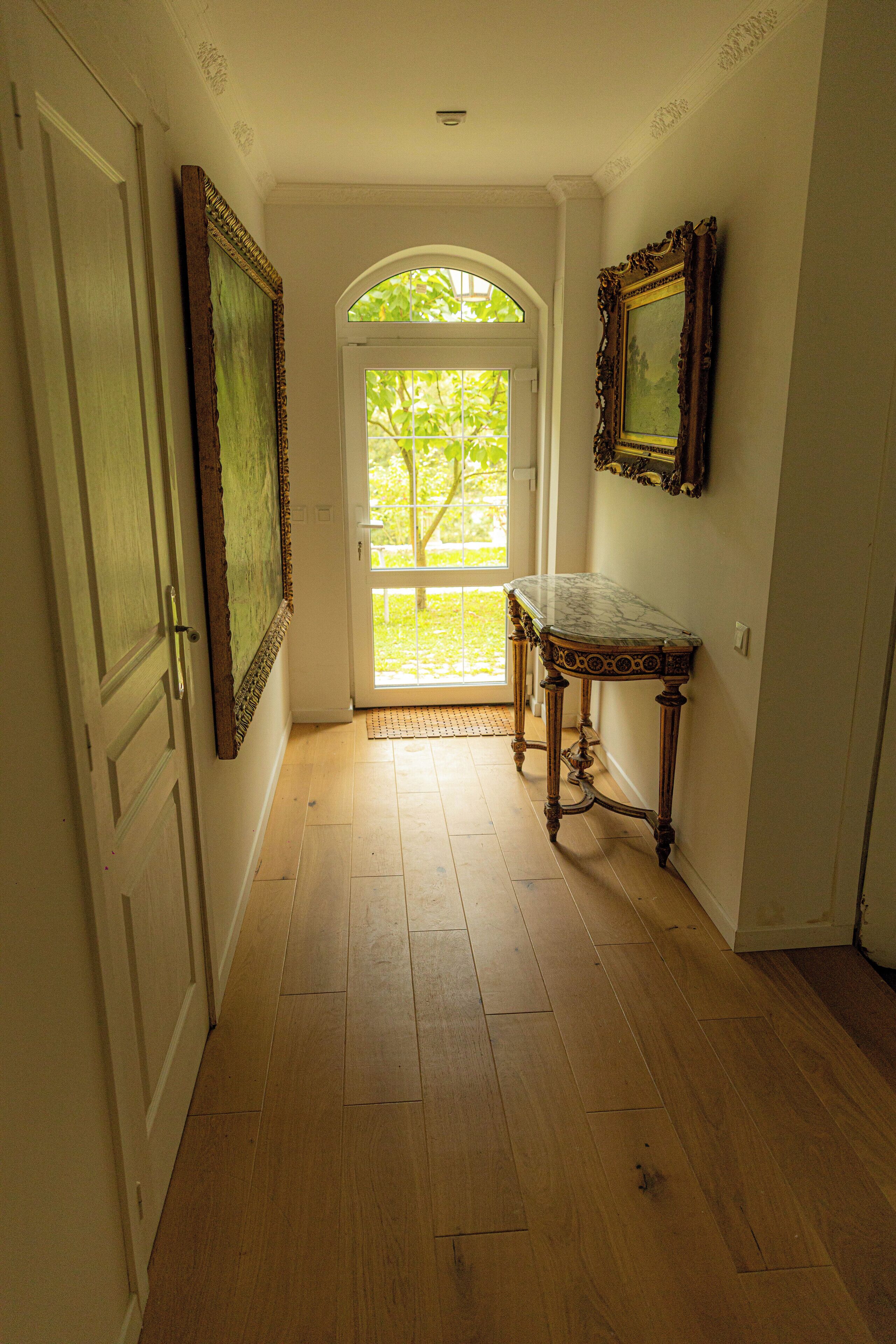 A hallway with wooden flooring, a door, a large painting, and a marble table.