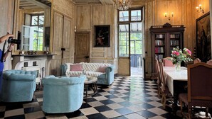 Um quarto com piso xadrez, painéis de madeira, um candelabro, lareira, estante de livros e uma mesa de jantar com cadeiras.