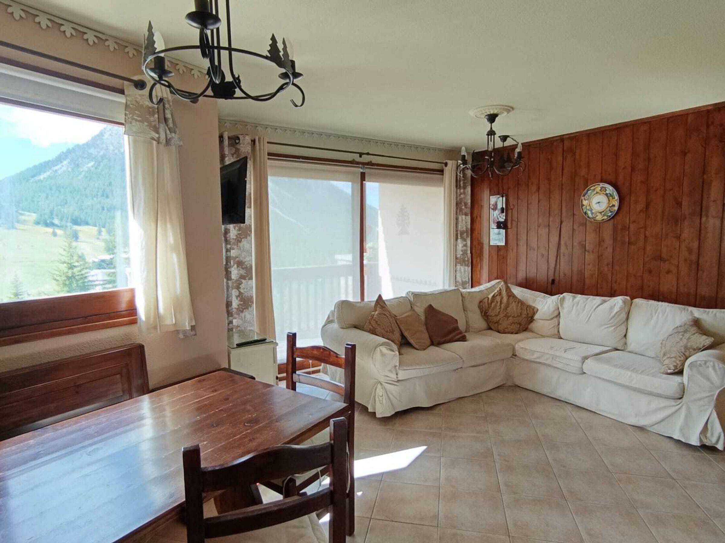 A living room with a wooden floor, a white sofa, a wooden dining table with chairs, and a view of mountains through a large window.