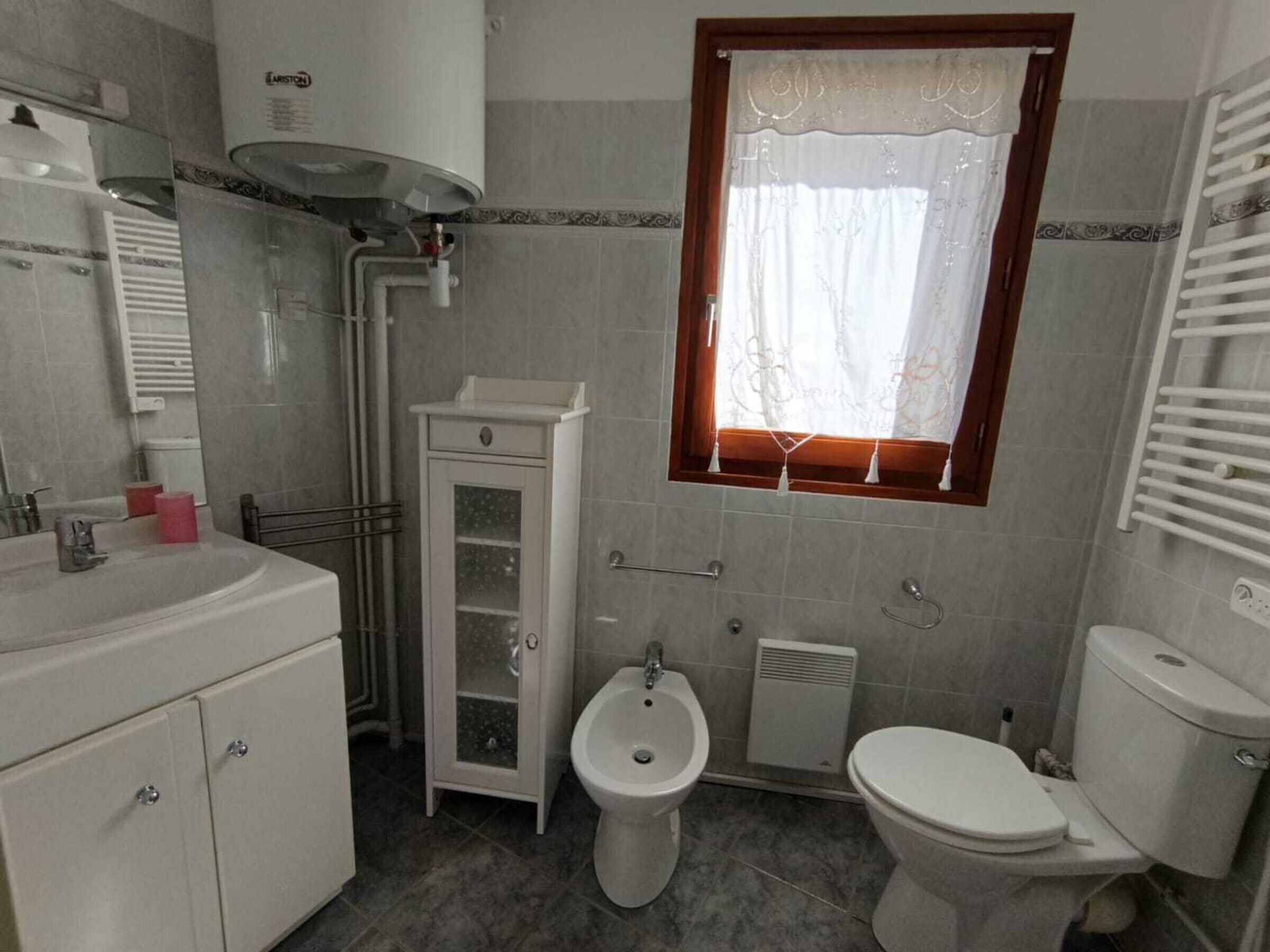 A bathroom with a toilet, bidet, vanity with cabinets, a window with curtains, and a radiator.
