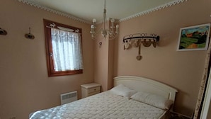 A bedroom with a bed, a nightstand, a heater, a window with curtains, a chandelier, and a wall-mounted shelf with a picture. - Functional apartment for 5 people with pets allowed, parking and south-facing balcony (Montgenèvre)