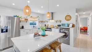 A modern kitchen with white cabinets, a stainless steel refrigerator, a washing machine, and a large island with a welcome sign. - BlueJay's Nest Unit B: Game Room, Walk to Water & Stadium (Clearwater)