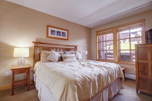 A bedroom with a large bed, wooden headboard, bedside tables, a lamp, and a window with a view of the outdoors. - Beautiful Condo in Passage Point! PP606 (Copper Mountain)
