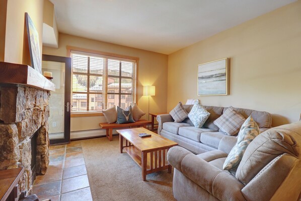 A living room with a stone fireplace, a sofa set, a wooden coffee table, and a large window with blinds. - Beautiful Condo in Passage Point! PP606 (Copper Mountain)