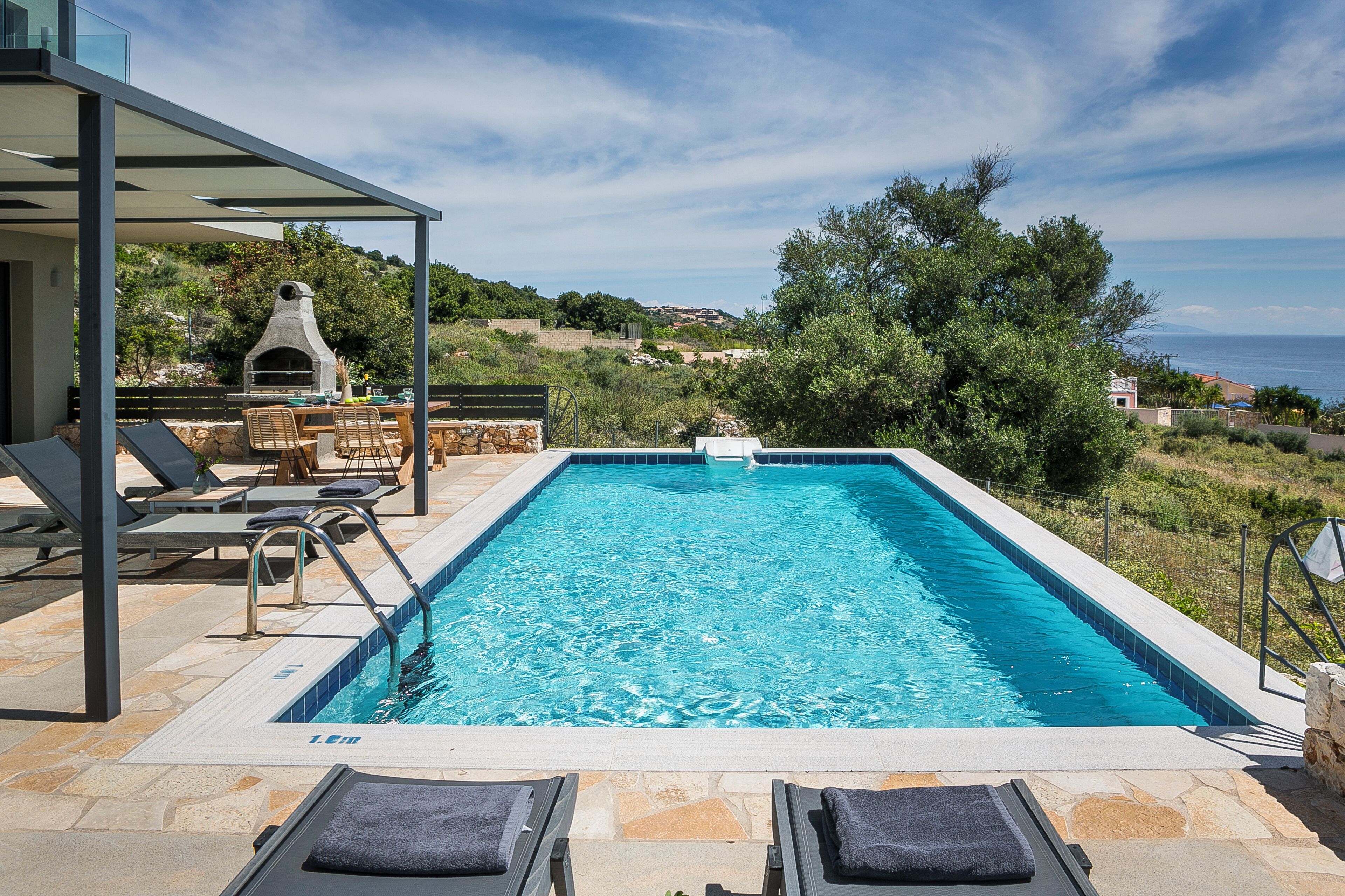 Una piscina rectangular con un borde de piedra, sillas de descanso y una parrilla de piedra.