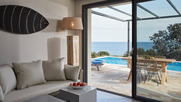 Ein modernes Wohnzimmer mit einer Couch, einem Tisch mit Obst, einer Lampe und Blick auf Pool und Meer durch große Glasfenster.