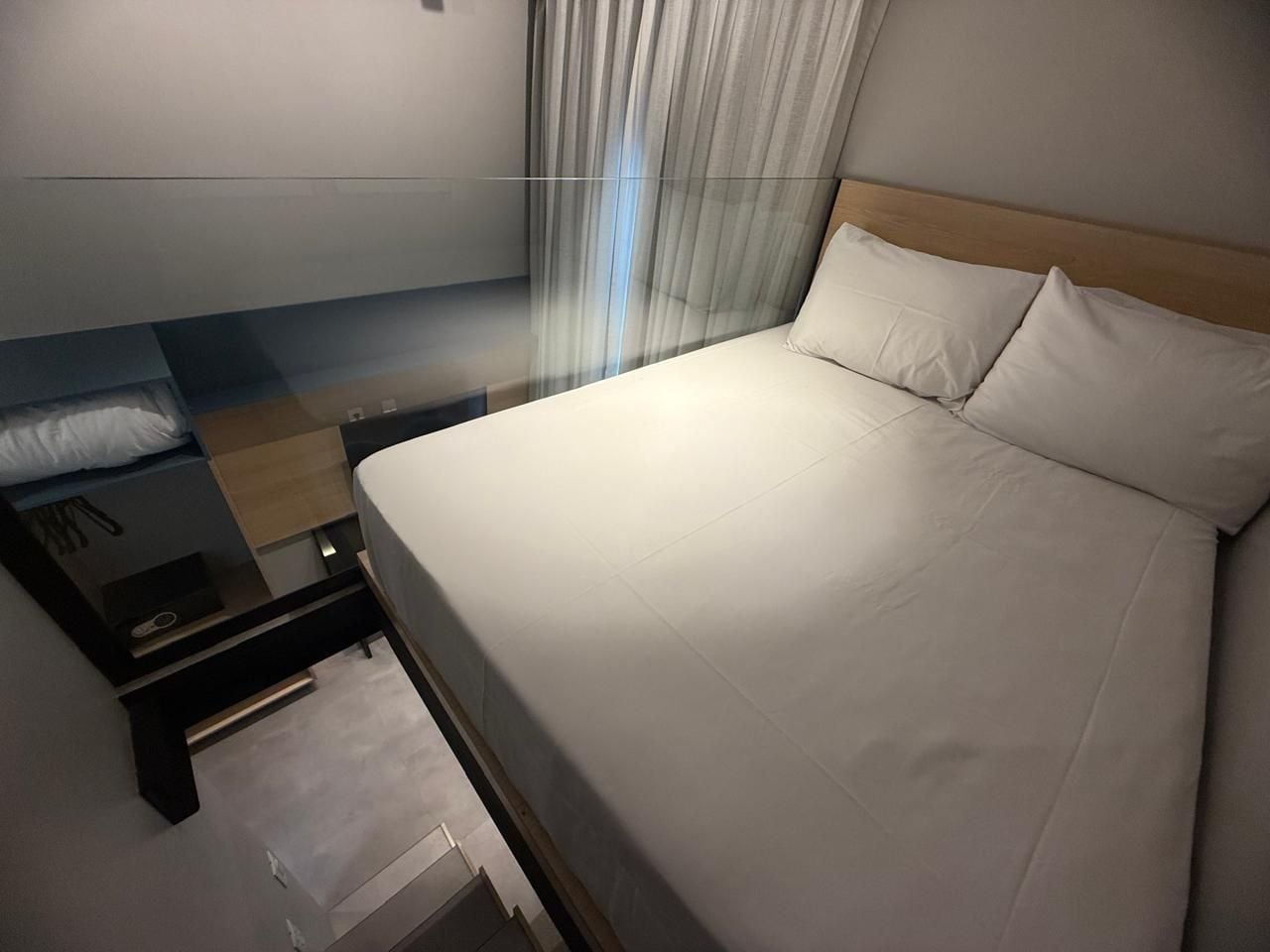 A hotel room with a bed, a nightstand, a drawer, a wooden headboard, and a window with curtains.