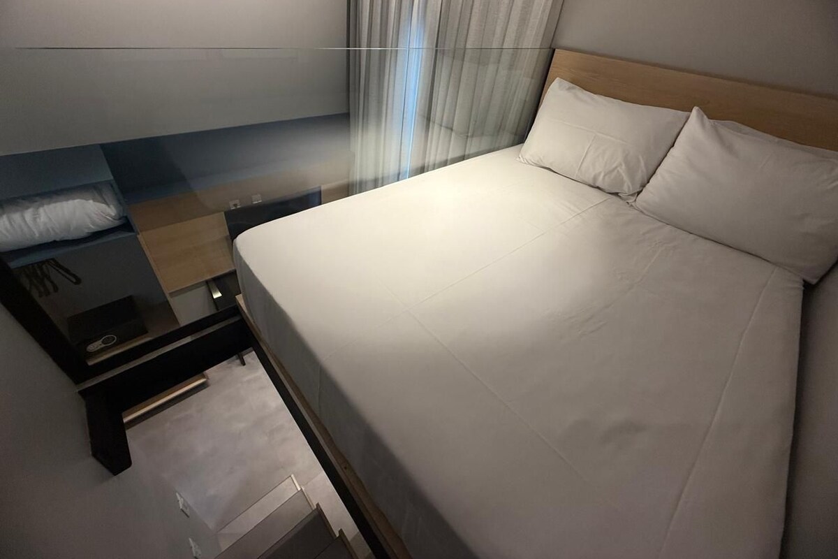 Hotelkamer met een bed, nachtkastje, lade, houten hoofdeinde en een raam met gordijnen.