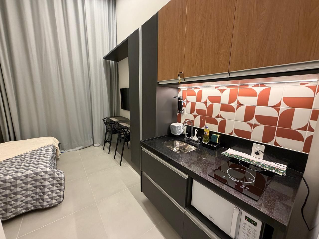 Habitación de hotel compacta con kitchenette, cama con cabecera de diseño geométrico, zona de comedor con una silla y un ventanal con cortinas.