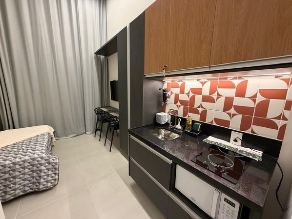 Habitación de hotel compacta con kitchenette, cama con cabecera de diseño geométrico, zona de comedor con una silla y un ventanal con cortinas.