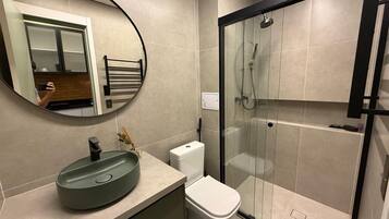 Un baño moderno con un espejo redondo, un lavabo, un inodoro y una zona de ducha con mampara de cristal.