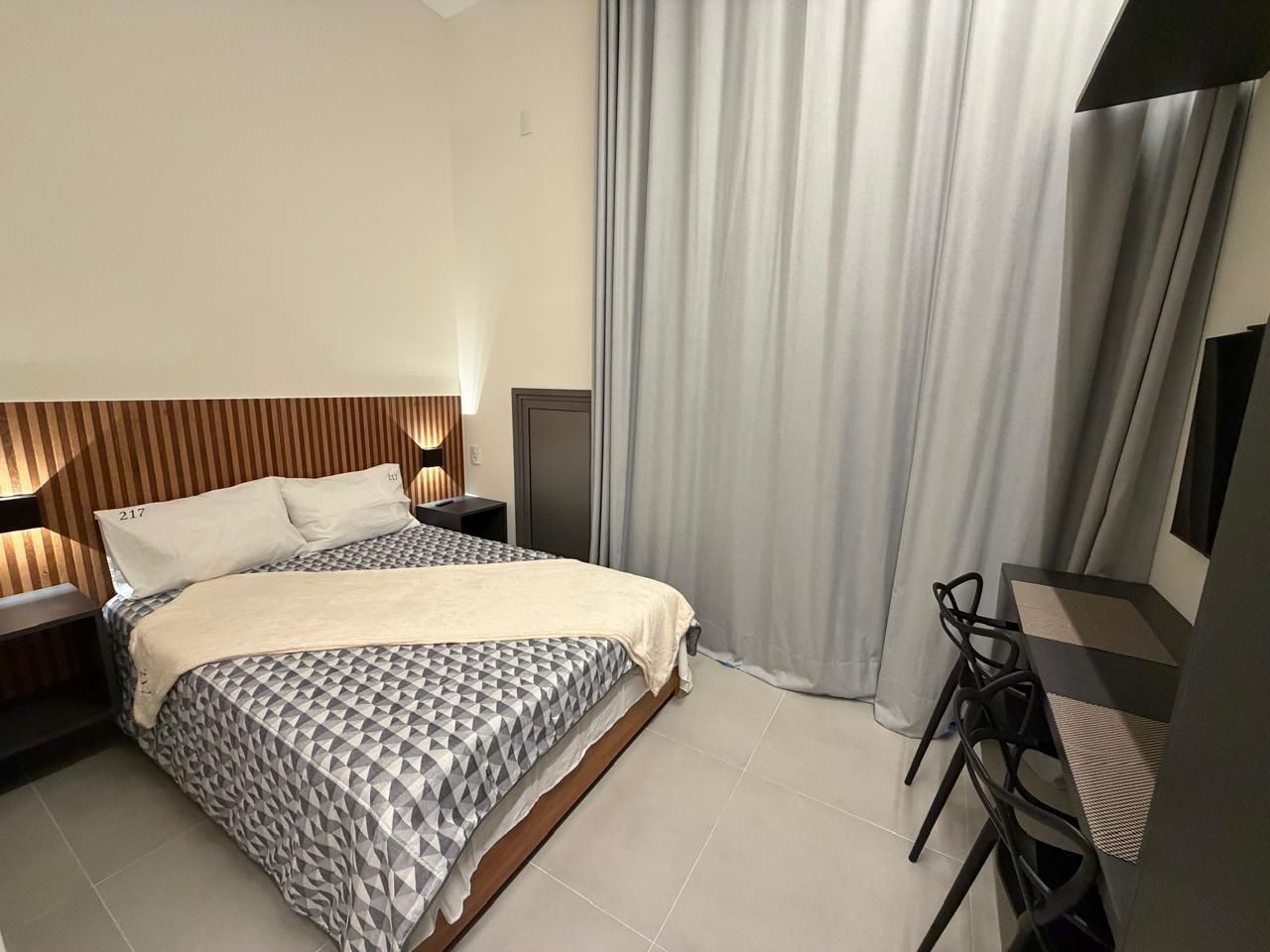 A modern hotel room with a bed, bedside table, lamp, and a desk with a chair.