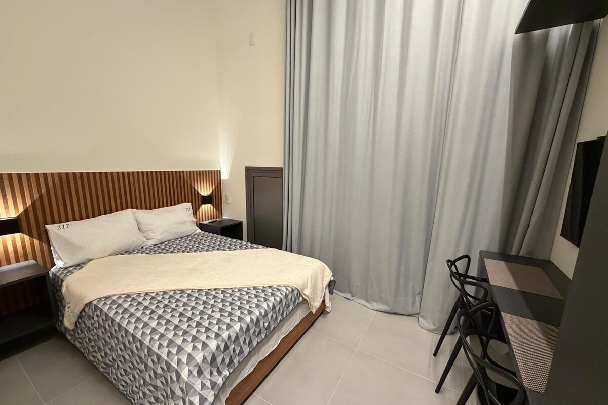 A modern hotel room with a bed, bedside table, lamp, and a desk with a chair.