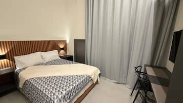 A modern hotel room with a bed, bedside table, lamp, and a desk with a chair.