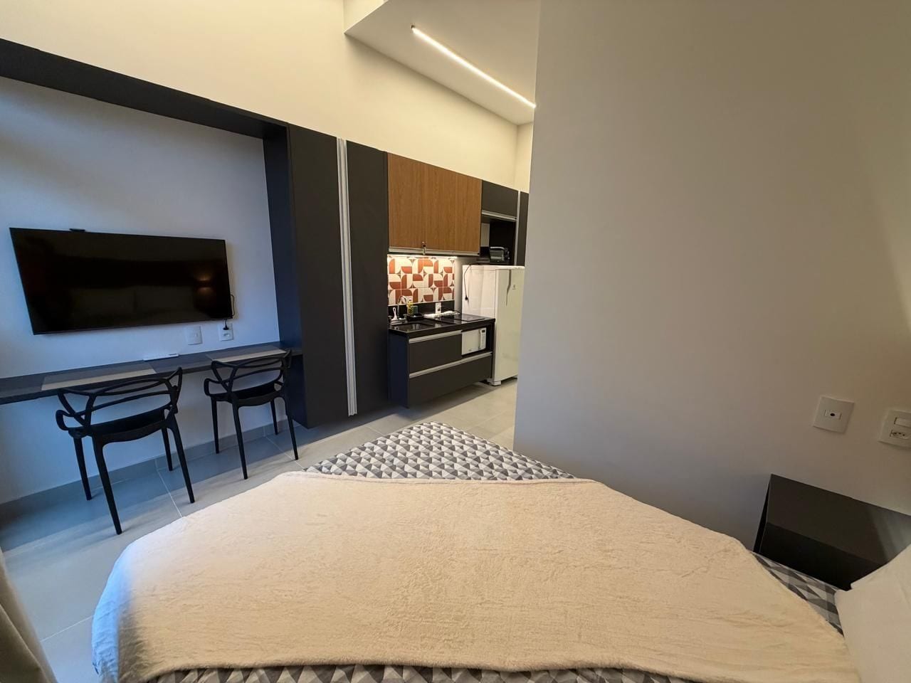A modern hotel room with a bed, a desk, two chairs, a TV, and a kitchenette in the background.