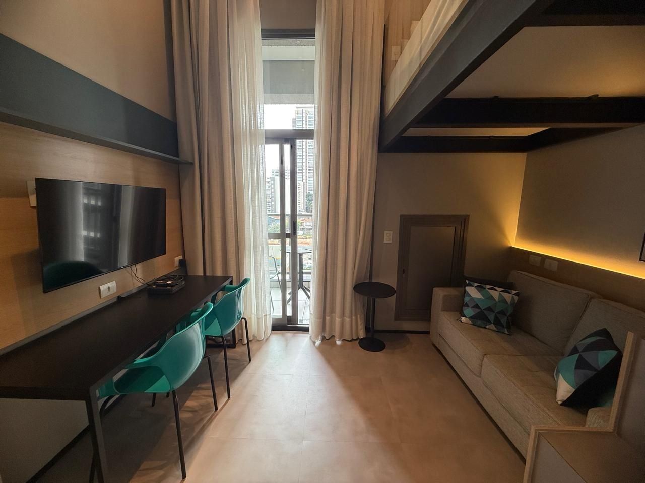 Ein kompaktes Hotelzimmer mit einem Flachbildfernseher, einem Esstisch mit Stühlen, einem Sofa mit Kissen und Blick durch ein großes Fenster.