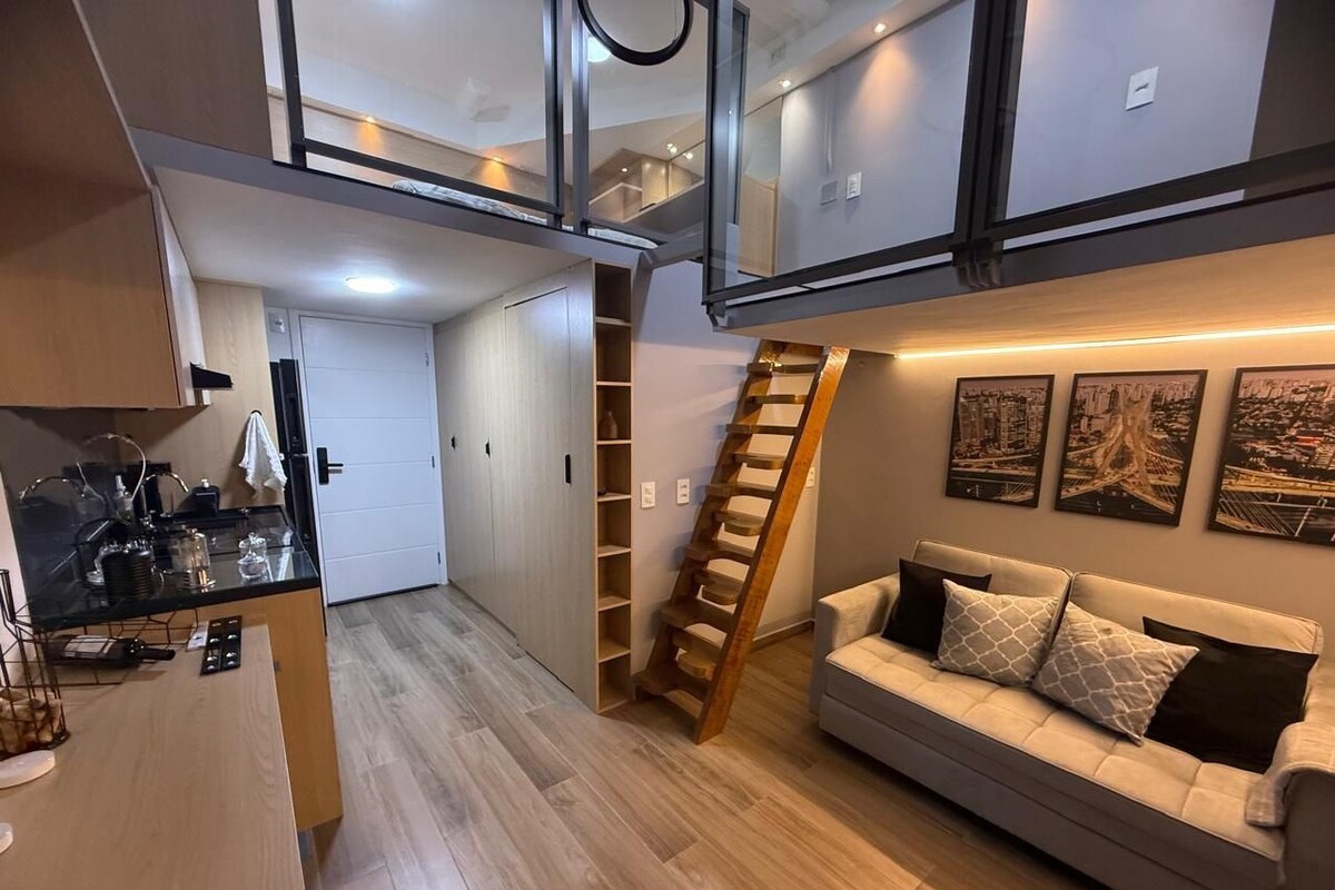 Un intérieur moderne comprenant une chambre à coucher aménagée en mezzanine, une cuisine et un espace de vie avec un canapé et des œuvres d’art.