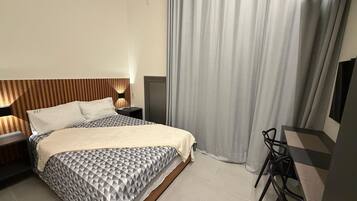 A modern hotel room with a bed, bedside table, lamp, and a desk with a chair.