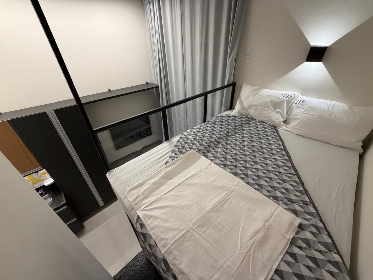Een enkel bed met een matras met geometrisch patroon en witte beddengoed, een zwart metalen hoofdbord, en een raam met gordijnen.