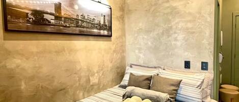 A hotel room with a bed, a cityscape painting, a wall-mounted light fixture, and a small stool.