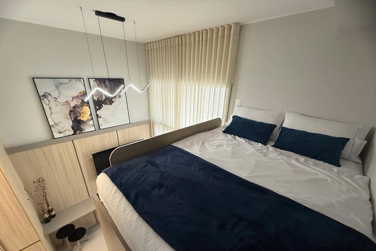 Une chambre à coucher moderne, avec un lit, des tables de chevet, un bureau et une grande fenêtre avec des rideaux.