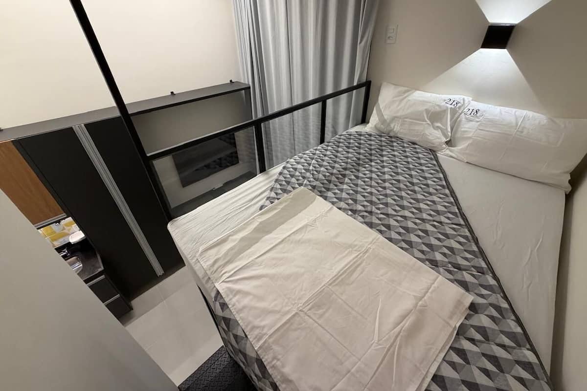 Un lit simple avec un matelas à motifs géométriques et du linge de lit blanc, une tête de lit en métal noir et une fenêtre avec des rideaux.