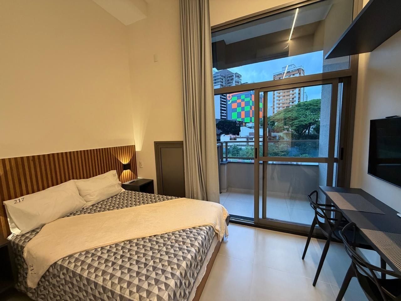 A modern hotel room with a large bed, a desk, and a balcony with a city view.