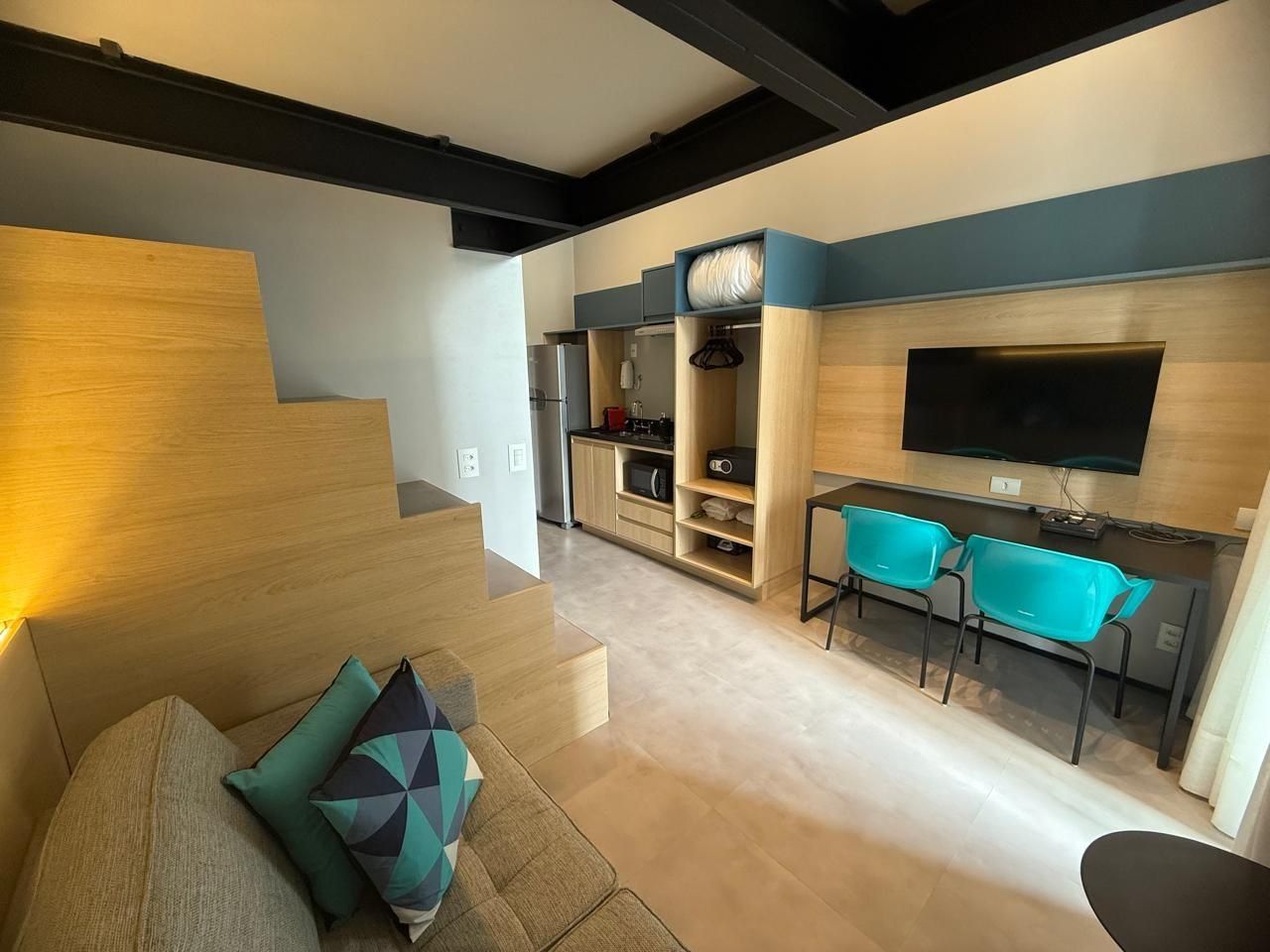 A modern living space with a staircase, a sofa with cushions, a dining area with chairs, a kitchen area with appliances, and a flat-screen TV mounted on the wall.
