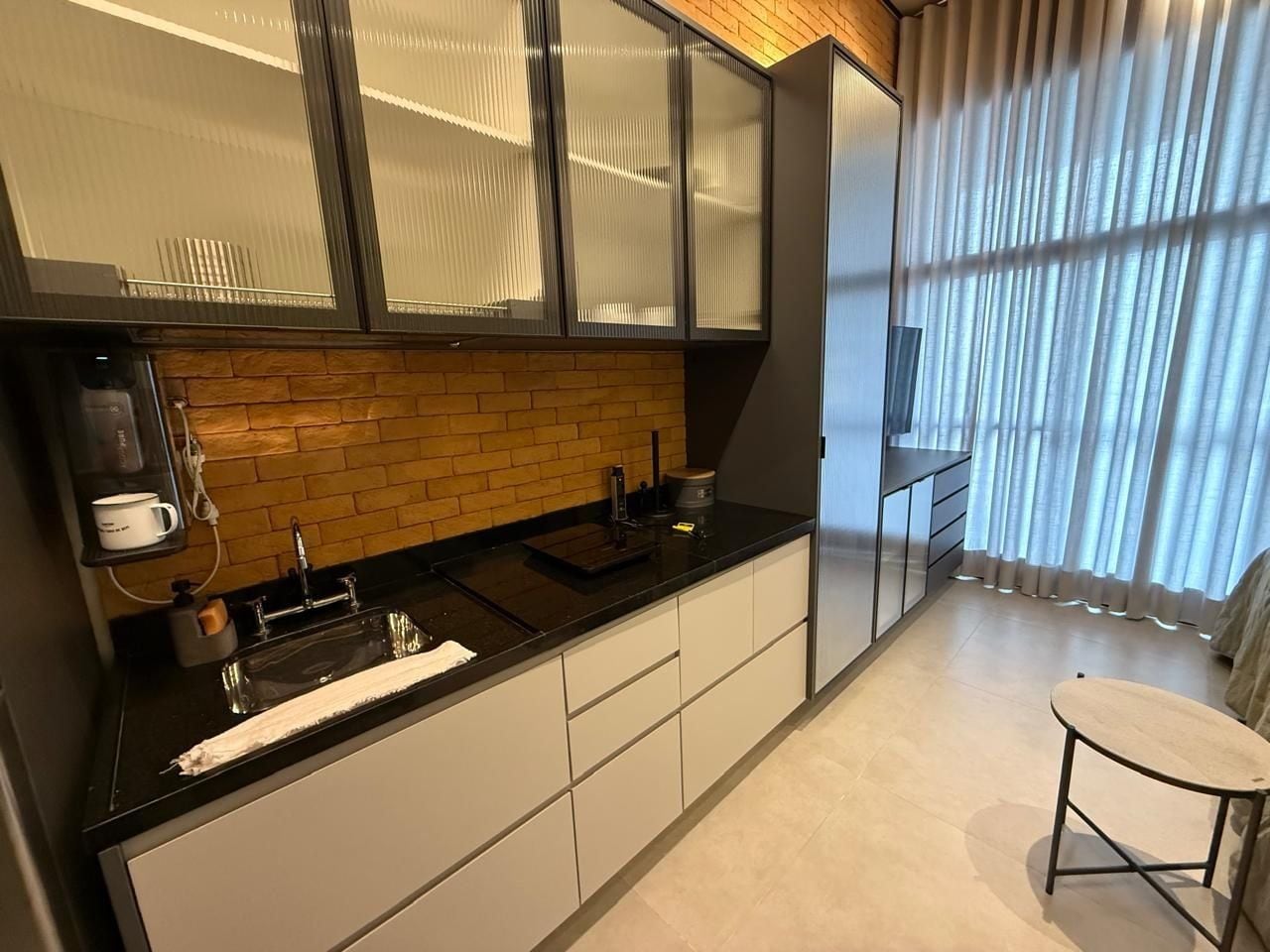 A modern kitchen with a black countertop, white cabinets, a brick backsplash, and a window with sheer curtains.