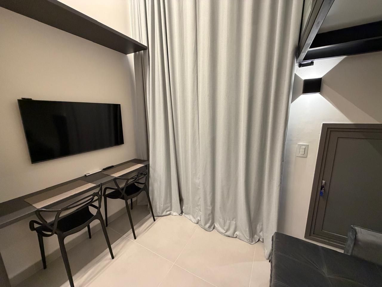 A modern room with a flat-screen TV mounted on the wall, a desk with two chairs, and a large window with curtains.