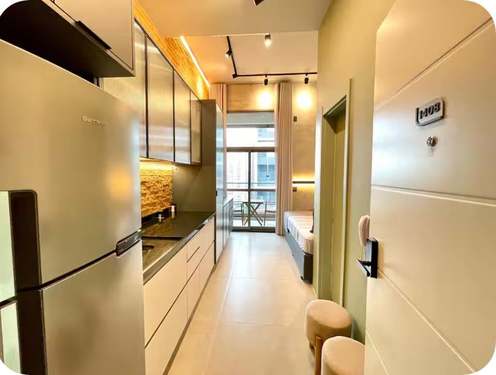A narrow hallway with a kitchenette on the left, a bed on the right, and a window at the end.