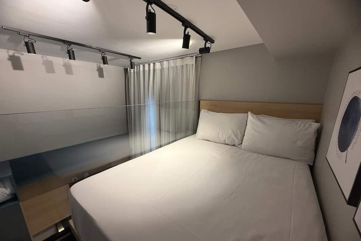 Une chambre d’hôtel comprenant un lit, un bureau avec une chaise, une fenêtre avec des rideaux et un luminaire fixé au plafond.
