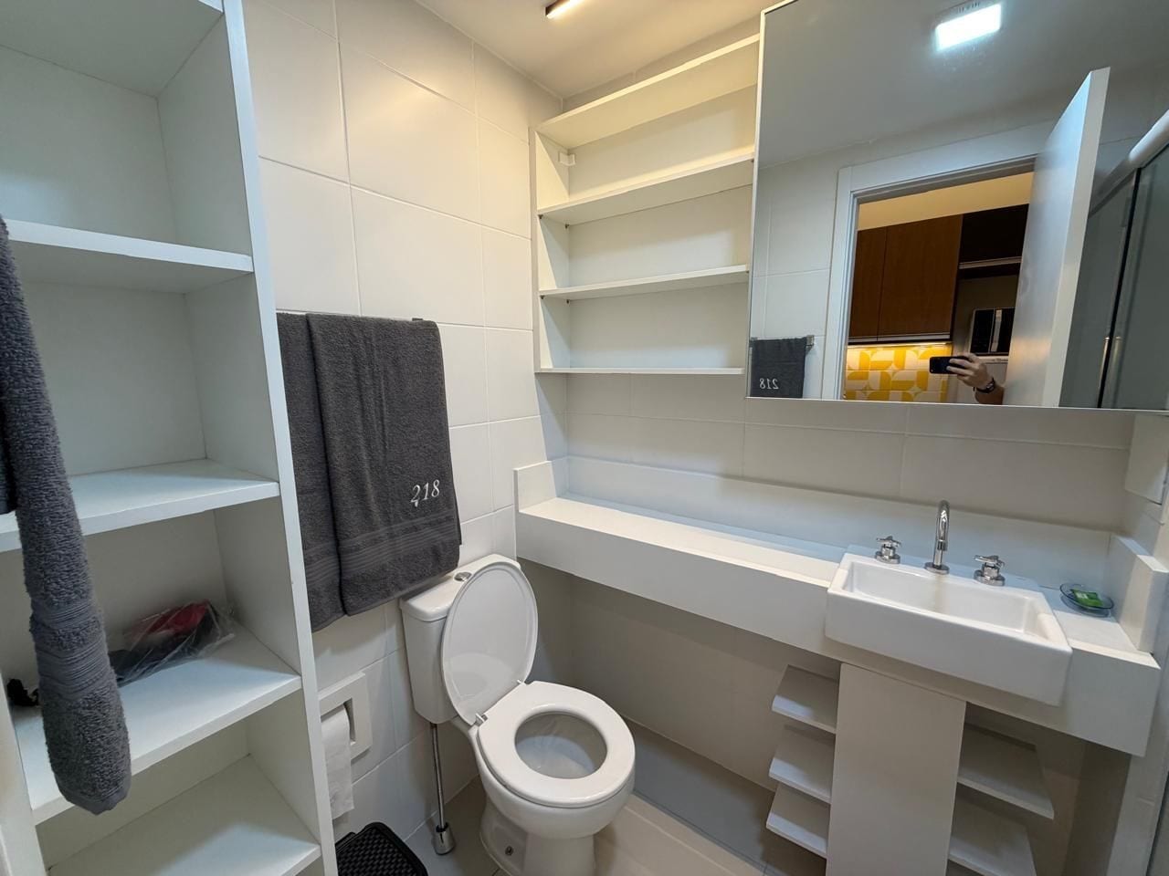 A bathroom with a toilet, a sink, and a mirror. There are shelves and a towel rack.