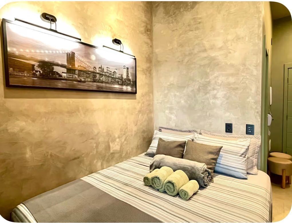 Une chambre d’hôtel avec un lit, un tableau représentant un paysage urbain, un luminaire fixé au mur et un petit tabouret.