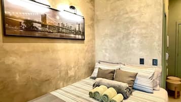 Une chambre d’hôtel avec un lit, un tableau représentant un paysage urbain, un luminaire fixé au mur et un petit tabouret.