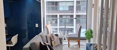 A modern living room with a grey sofa, a glass coffee table, and a view of a building through large windows.