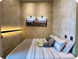 Een compacte hotelkamer met een bed, een aan de muur bevestigd schilderij en een lichtbalk langs de muur.