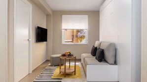 A compact living space with a sofa, a small table with fruit, a television, and a window with a view of the outside. - Waou Seville Center I (Sevilla)