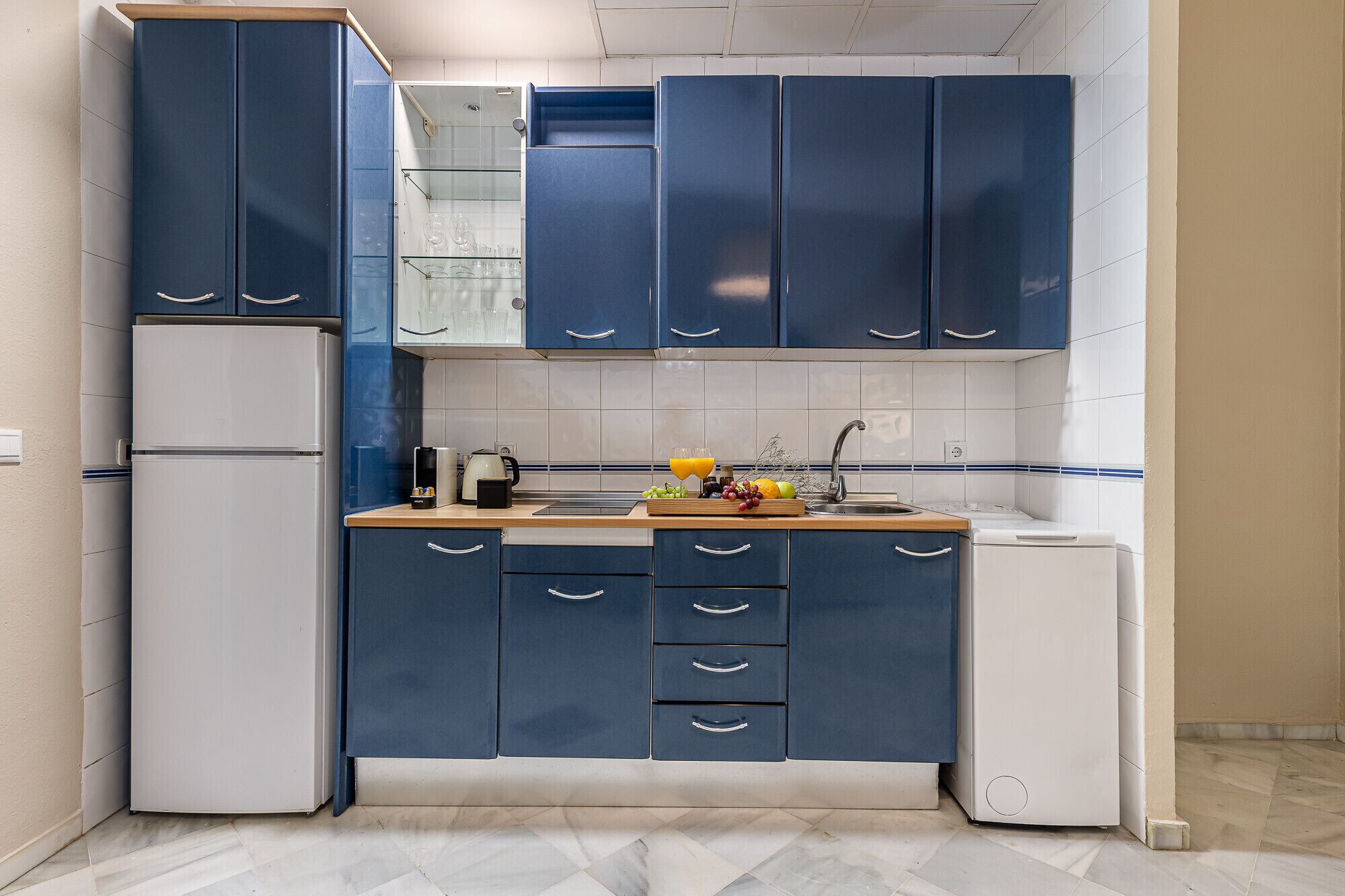A compact kitchen with blue cabinets, a white refrigerator, a sink, and a small washing machine.