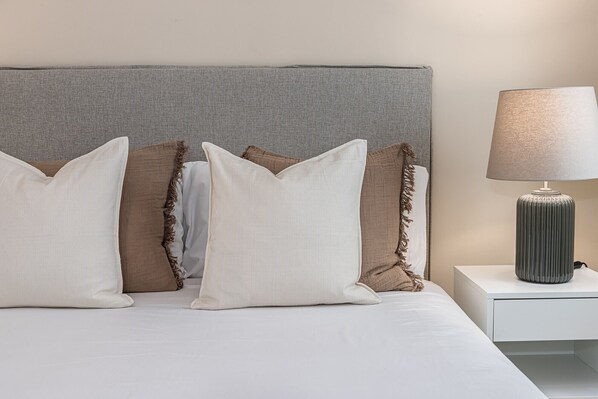 A bed with white bedding, a grey headboard, and three pillows in white, brown, and beige. A lamp with a ribbed base is on a bedside table. - Waou Seville Center III (Sevilla)