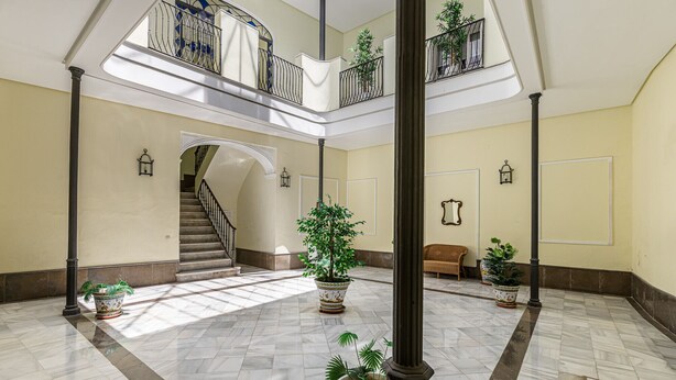 A spacious, well-lit atrium with a marble floor, multiple potted plants, a staircase leading to upper levels, and a wicker sofa. - Waou Seville Center III (Sevilla)